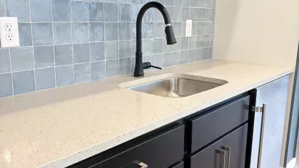 Modern kitchenette with a speckled light countertop, black cabinets, matte black faucet, and glossy blue tile backsplash beneath two floating shelves.