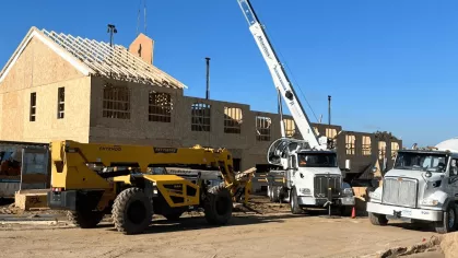A large crane lifts a prefabricated wall panel into place next to a yellow construction vehicle at the active building site.
