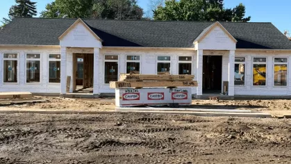Exterior view of a single-story building under construction with installed windows, Tyvek wrap, and stacked lumber in the foreground.