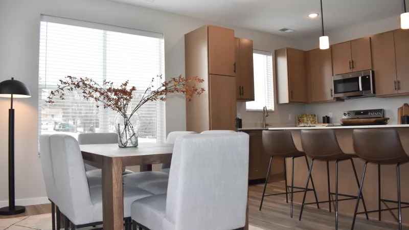 Modern open-concept dining area with wood table, grey chairs, and a kitchen featuring wood cabinets and island with brown stools.