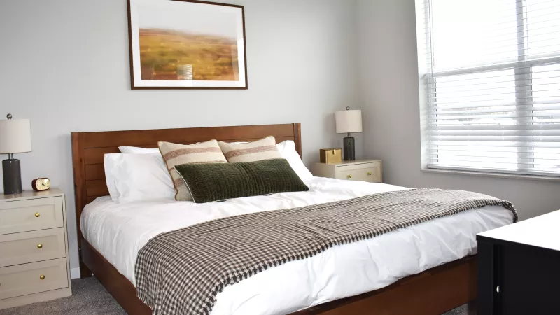Modern bedroom with a queen bed, wooden frame, white bedding, nightstands with lamps, and a landscape painting.