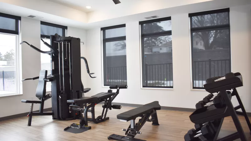 Well-lit modern gym with a multi-station machine, weight benches, and dumbbells on a wooden floor, white walls, and dark blinds.