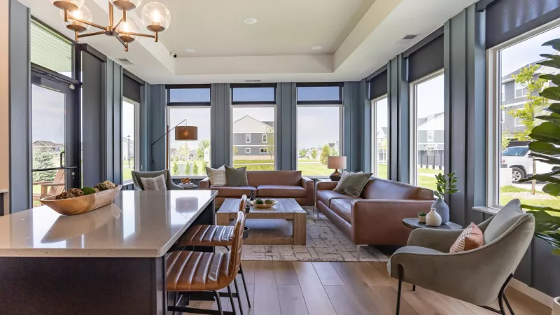 Modern living room with brown leather sofas, a grey chair, and large windows overlooking a neighborhood. Kitchen island foreground.