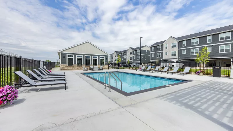 Modern outdoor swimming pool, lounge chairs, a clubhouse, and gray apartment buildings under a partly cloudy sky.