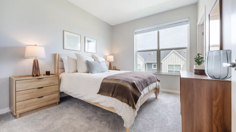 Bright, modern bedroom featuring a queen bed with wood frame, light gray walls and carpet, and a dark wood dresser.