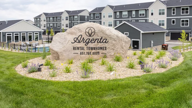 Large rock sign for Argenta Rental Townhomes, with modern apartment buildings and a pool behind it.