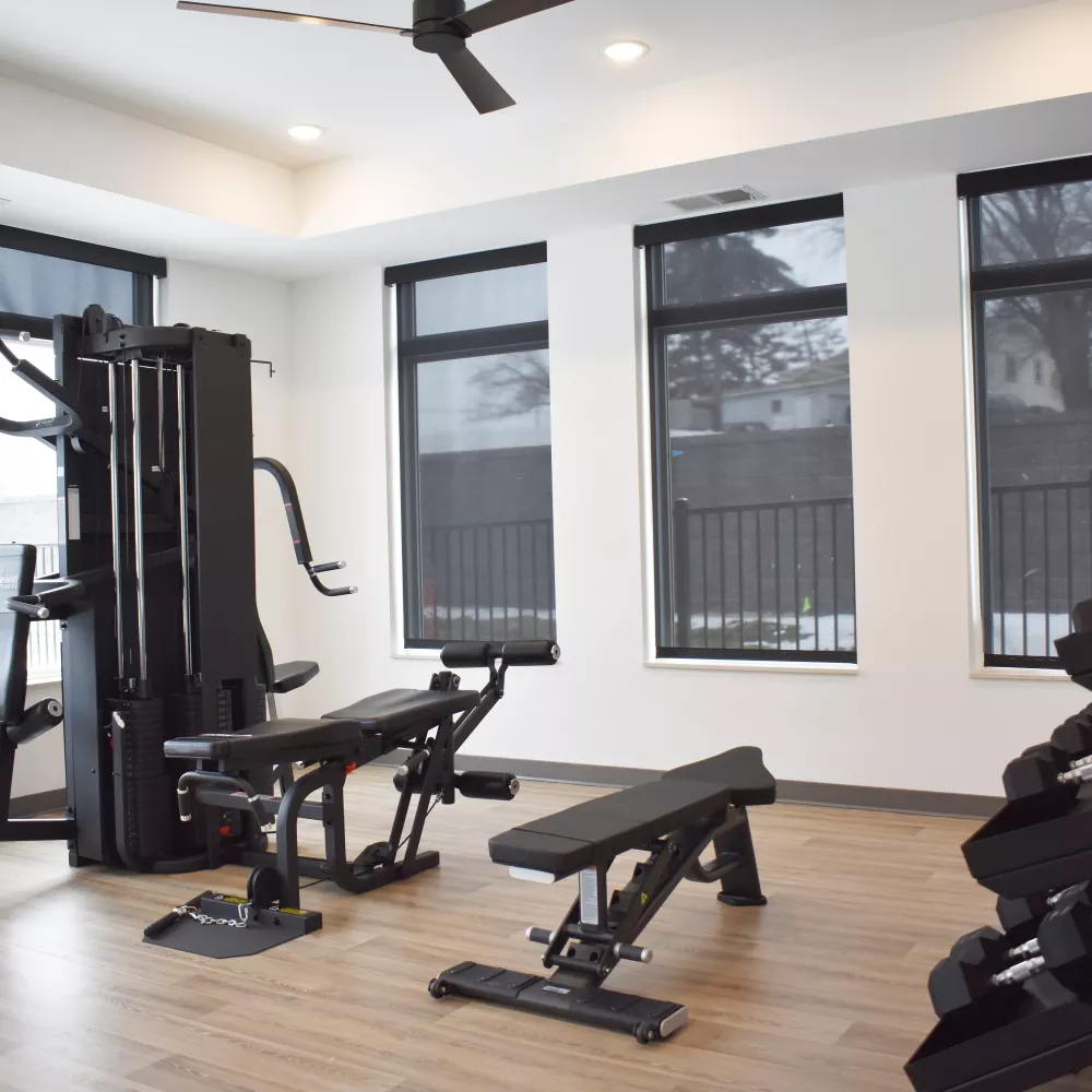 Well-lit modern gym with a multi-station machine, weight benches, and dumbbells on a wooden floor, white walls, and dark blinds.