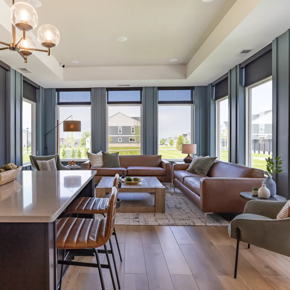 Modern living room with brown leather sofas, a grey chair, and large windows overlooking a neighborhood. Kitchen island foreground.