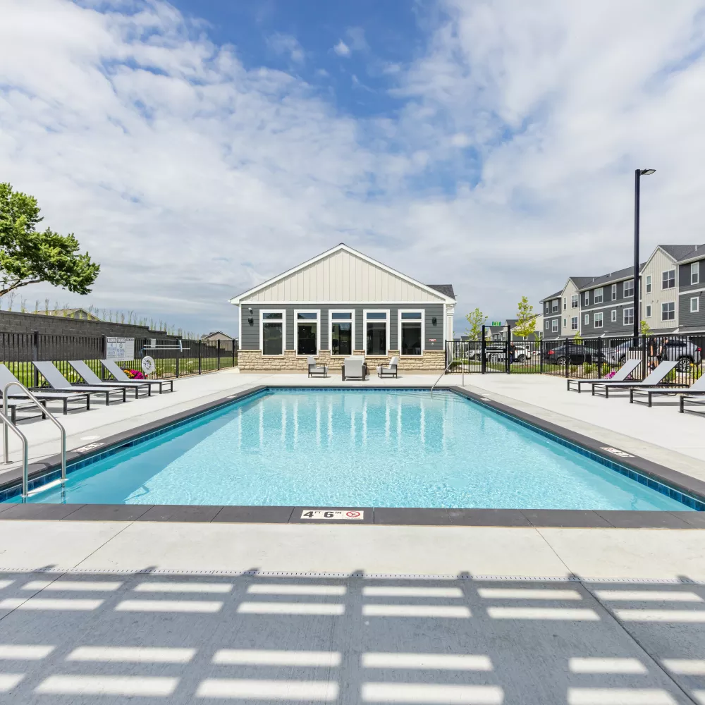 Outdoor swimming pool with lounge chairs, a clubhouse, and apartment buildings under a partly cloudy sky.