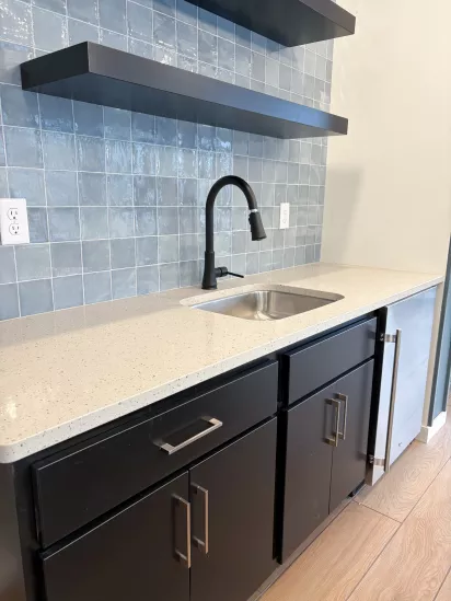 Modern kitchenette with a speckled light countertop, black cabinets, matte black faucet, and glossy blue tile backsplash beneath two floating shelves.