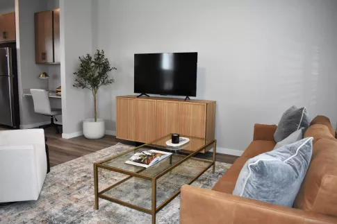 Stylish living room featuring a brown couch, glass coffee table, flat-screen TV, and a glimpse of a kitchen.