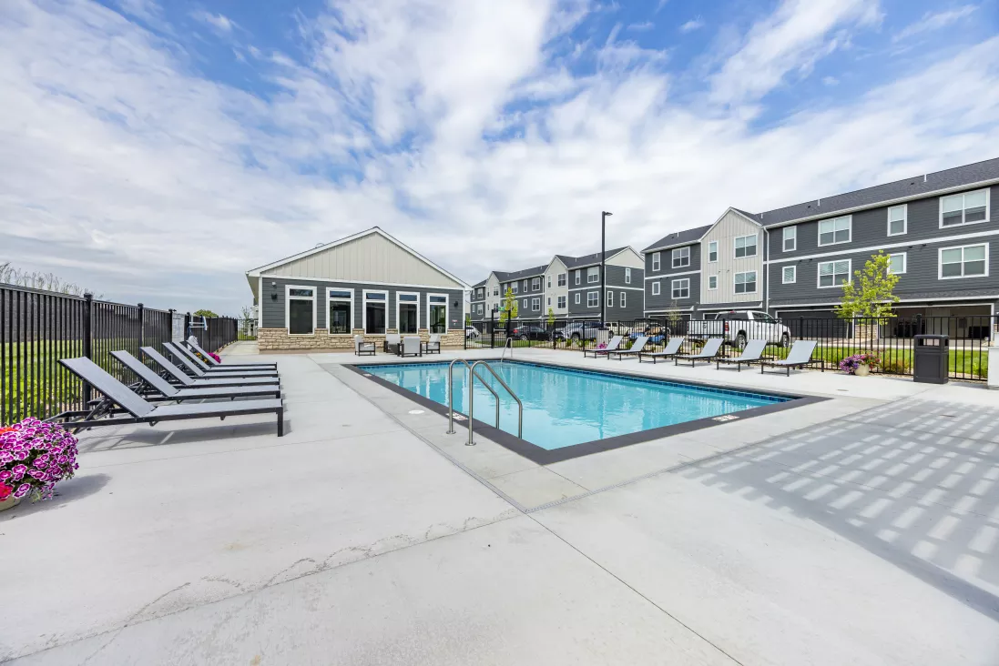Modern outdoor swimming pool, lounge chairs, a clubhouse, and gray apartment buildings under a partly cloudy sky.