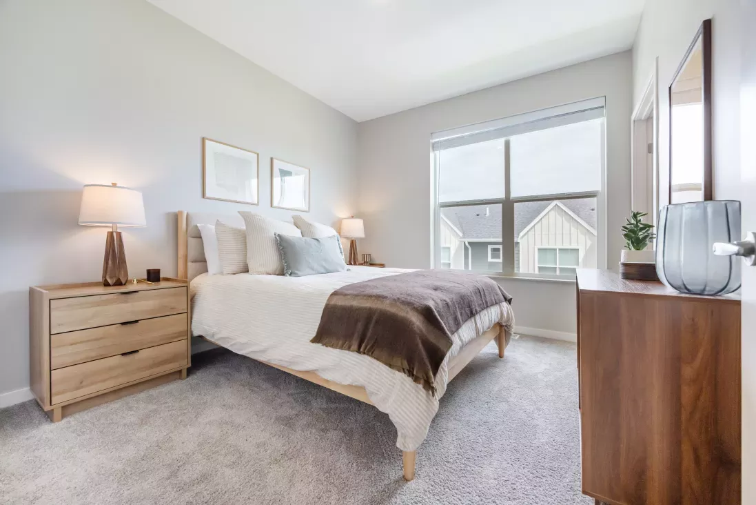 Bright, modern bedroom featuring a queen bed with wood frame, light gray walls and carpet, and a dark wood dresser.