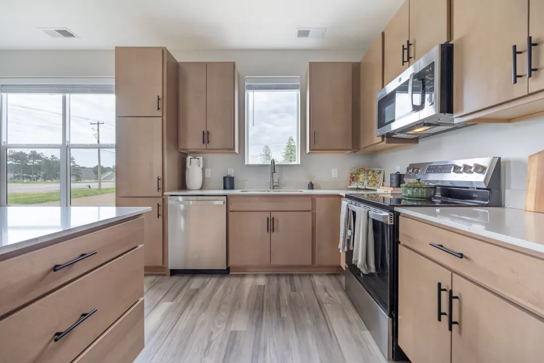 Modern kitchen with light wood cabinets, stainless steel appliances, white countertops, and wood-look flooring.